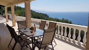 A covered outdoor dining area with a table and chairs, overlooking a scenic view of the sea and hills. - Villa Theodora in Nisaki (Corfu)