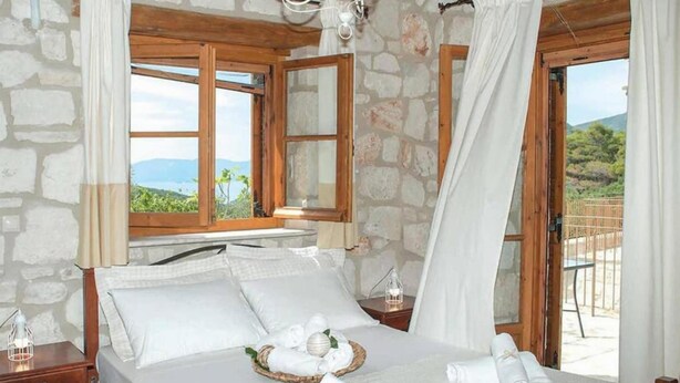A bedroom with a stone wall, wooden beams, a bed with white linens, a bedside table with a lamp, and a view of the outdoors. - Villa Aloni in Koridallos (Zakynthos)