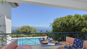 Un balcón con piscina, dos sillas rosadas con cojines, una mesa con platos de comida y vistas al mar y a las montañas.