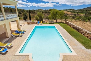 A rectangular swimming pool with a clear blue water, surrounded by a stone deck. There are lounge chairs with blue and yellow cushions arranged around the pool.