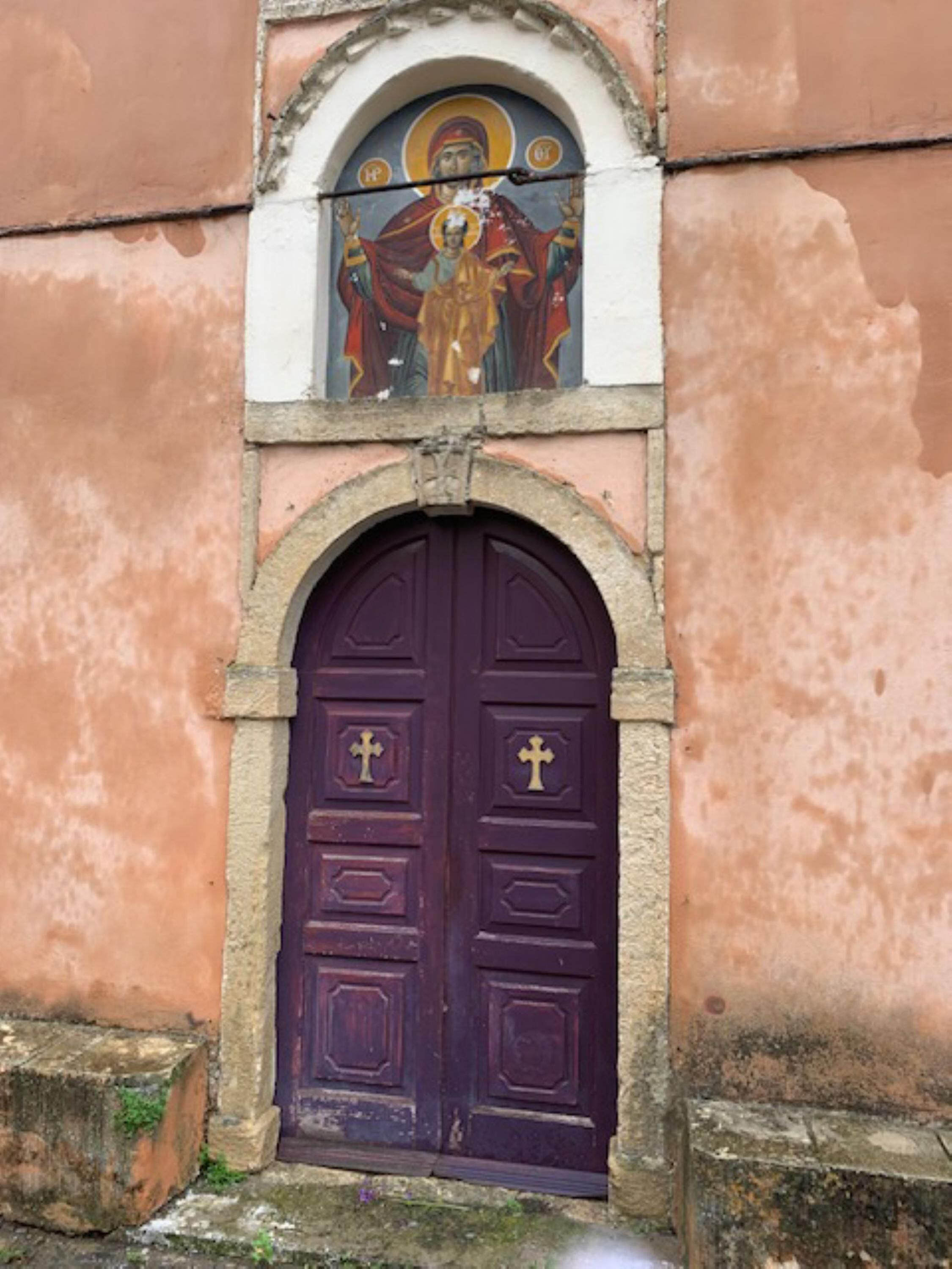 A purple door with a cross symbol, set within a stone archway, above a pink wall.