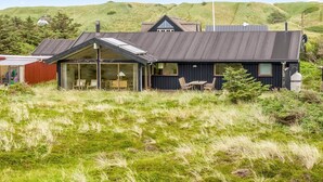 Ein modernes Haus mit einer großen Glasfront, einem Essbereich mit Außenbestuhlung und Blick auf sanfte Hügel im Hintergrund.
