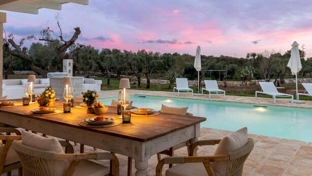 A wooden dining table with chairs, a pool, and lounge chairs in the background. - Villa Belkis 6 Emma Villas (Ostuni)