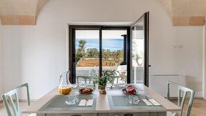 A dining area with a table set for two, a view of the outdoors, and a small kitchenette. - Villa Belkis 6 Emma Villas (Ostuni)