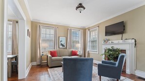 A living room with a beige sofa, two blue armchairs, a fireplace, and a flat-screen TV mounted on the wall. - Pool & Mins to Zoo - Capitol Heights Hideaway (Montgomery)