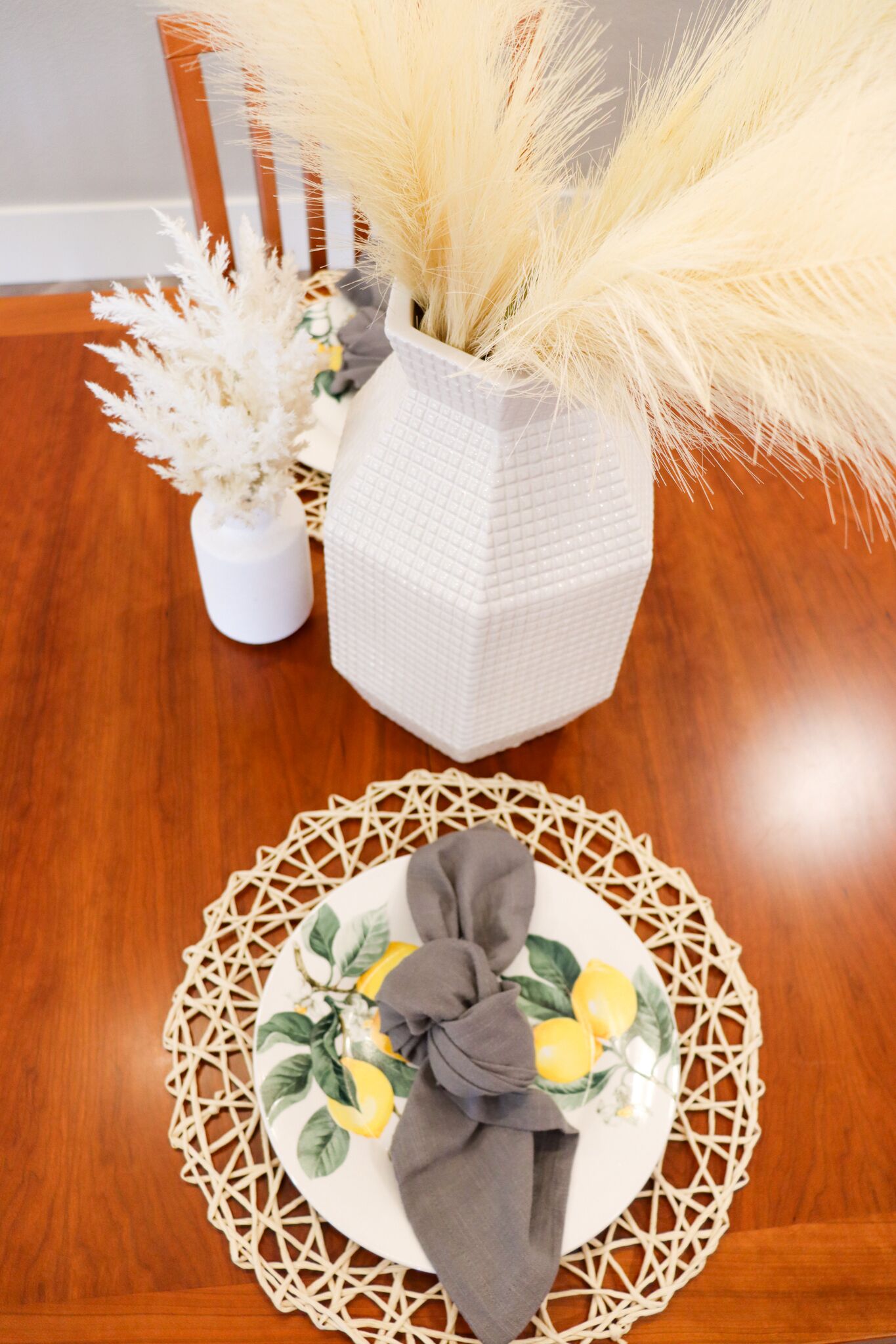 Uma mesa de jantar de madeira com um vaso branco contendo plumas de capim-do-campo e outro vaso de cerâmica branca com flores secas. Um jogo americano trançado com um padrão de limão está sobre a mesa.