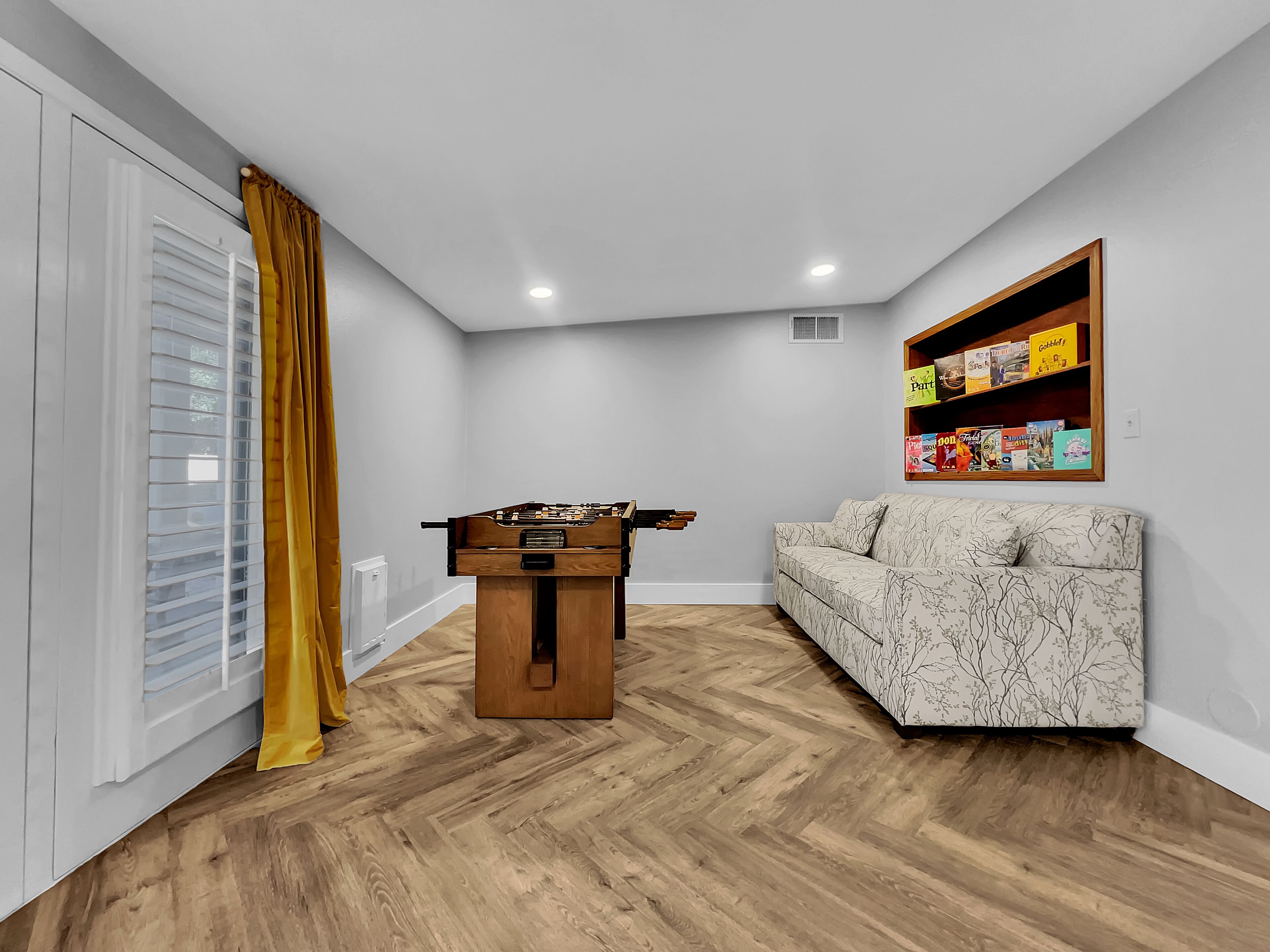 A room with a wooden floor, a sofa, a foosball table, a window with blinds, and a shelf with books.