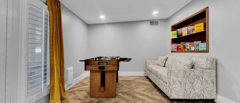 A room with a wooden floor, a sofa, a foosball table, a window with blinds, and a shelf with books.