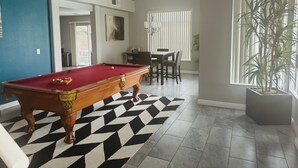 A pool table in a room with a geometric patterned rug, a dining area with chairs, a potted plant, and a window with blinds. - Relax with the whole family at this peaceful place to stay. (North Las Vegas)