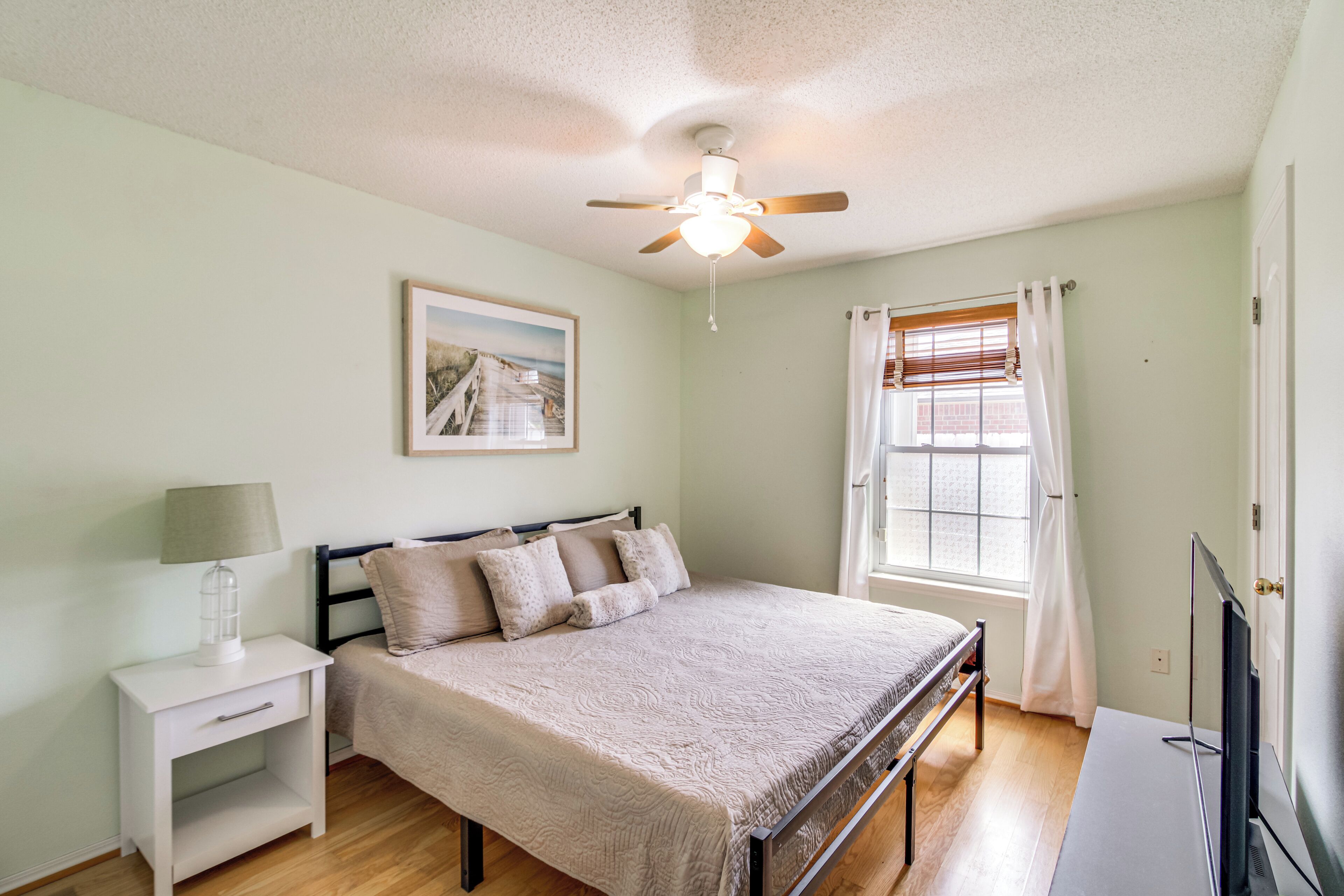 A bedroom with a bed, nightstand, ceiling fan, window with curtains, and a TV.