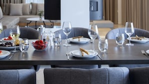 Un coin repas avec une table dressée pour quatre personnes, entouré de chaises à revêtement gris. Un téléviseur à écran plat est fixé au mur, et la pièce est dotée de grandes fenêtres avec des rideaux.