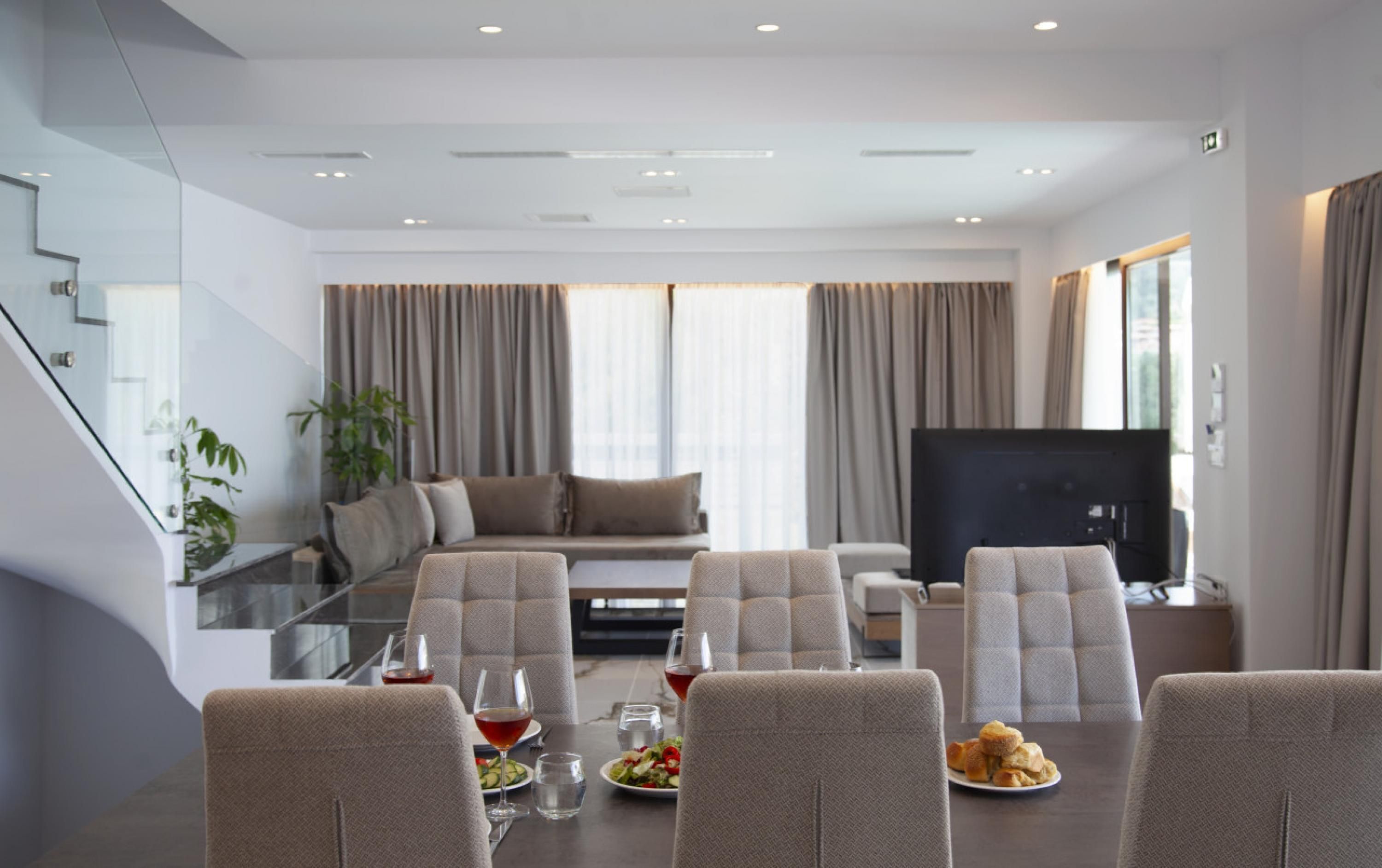 A modern living room with a glass dining table, comfortable seating, a flat-screen TV, and large windows with curtains.