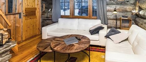 A log cabin interior with a white sofa, two round wooden tables, a patterned rug, and a stone fireplace.