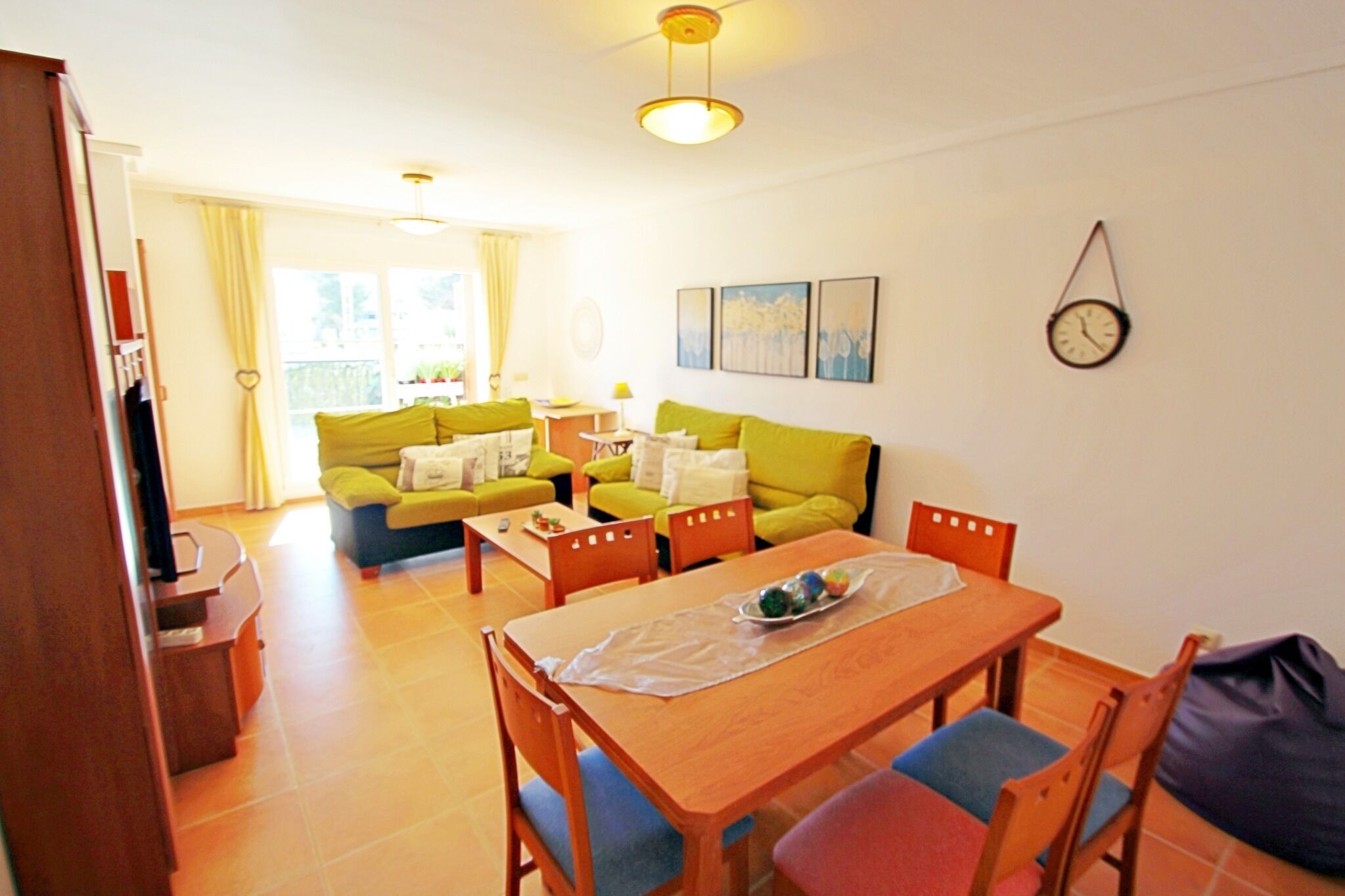 A living room with a wooden dining table, green sofa, and a clock on the wall.