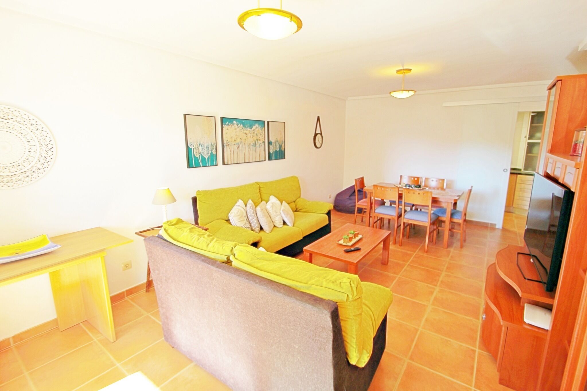 Uma sala de estar com sofá amarelo, mesa de jantar com cadeiras, rack para TV e obras de arte na parede.