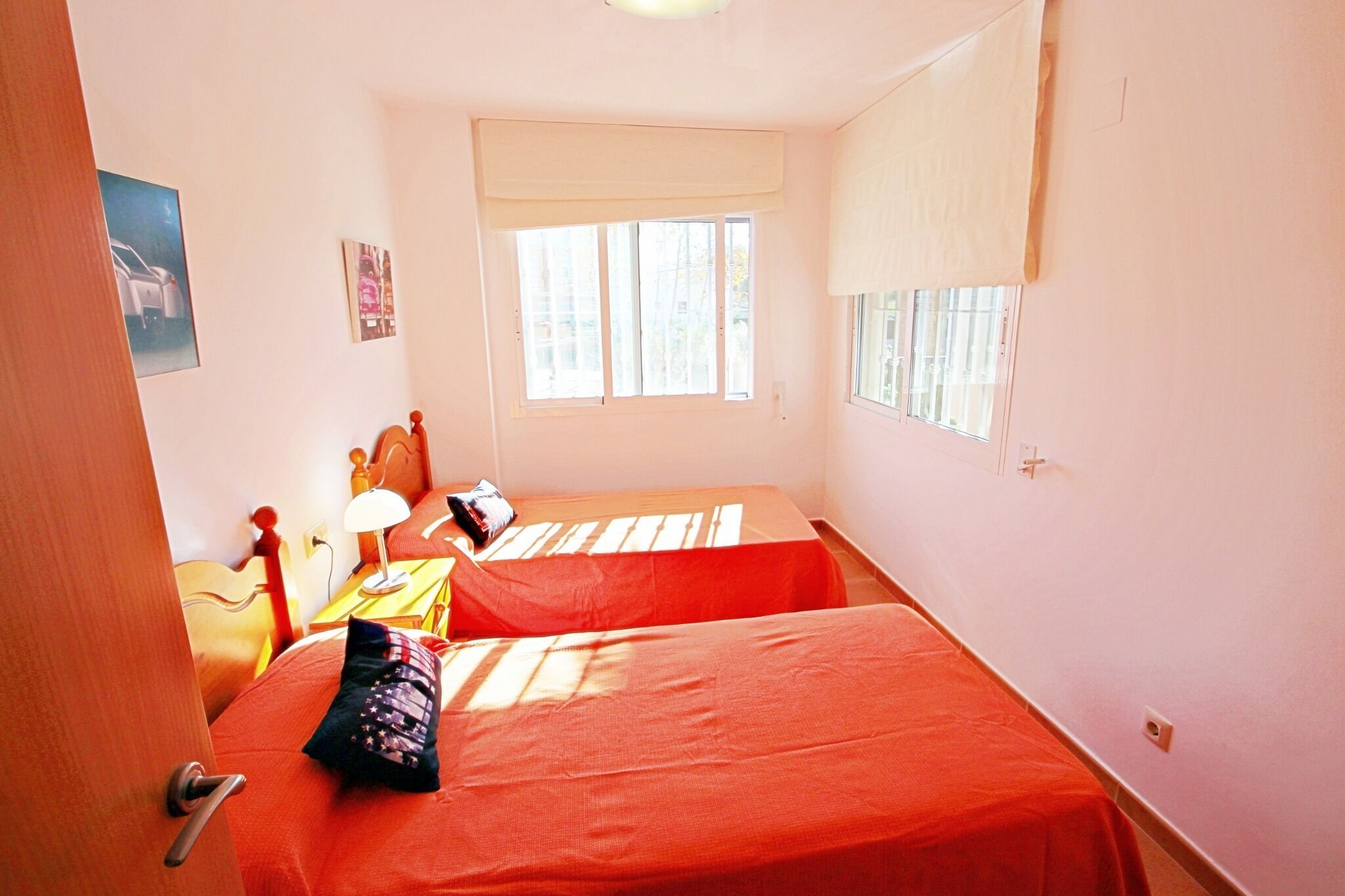 A room with two beds, a wooden headboard, a desk with a lamp, and a window with blinds.