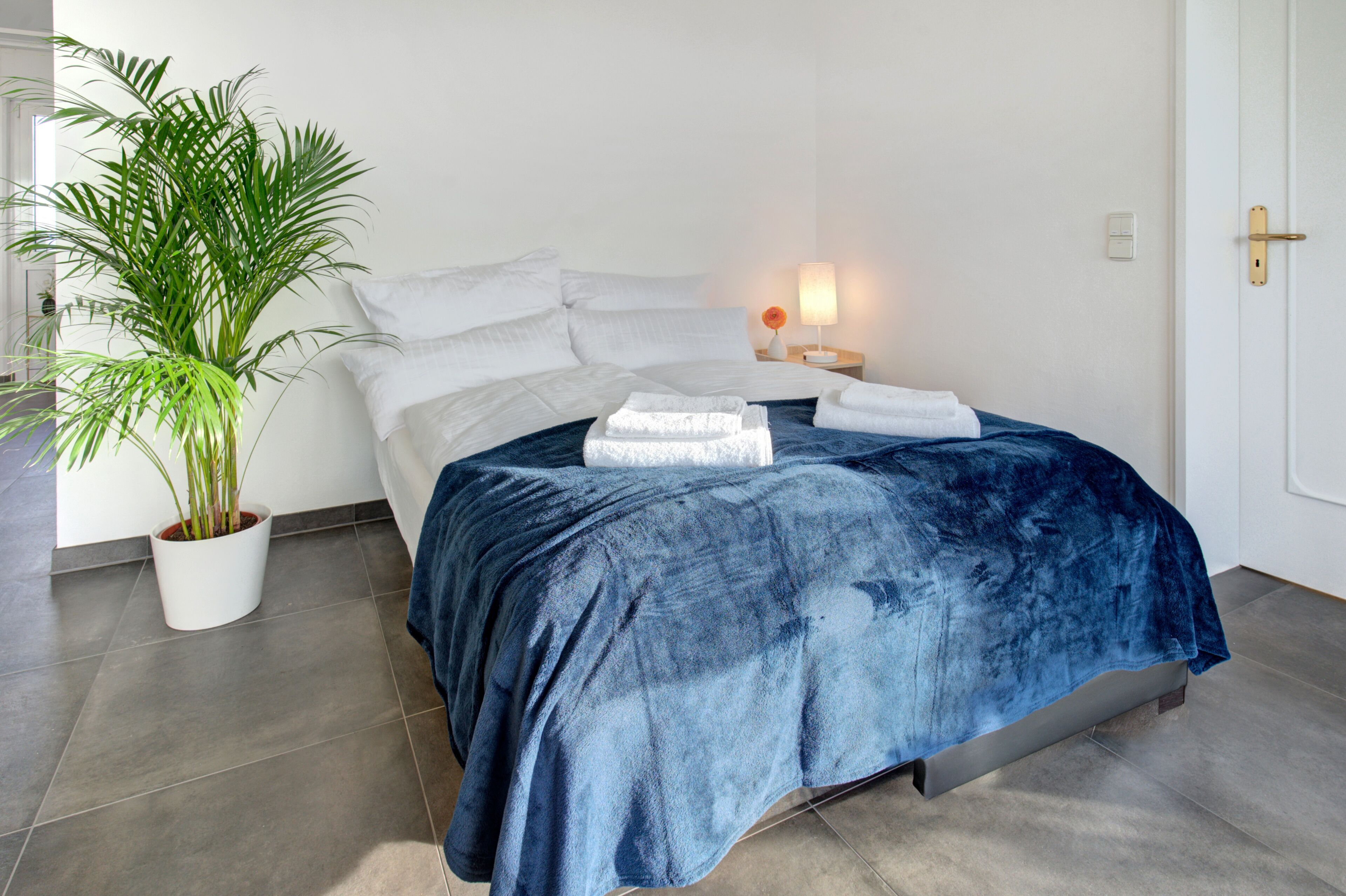 Ein ordentlich gemachtes Bett mit blauer Decke, einem Blumentopf und einer Nachttischlampe.
