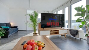 A modern living room with a flat-screen TV displaying the Disney+ Netflix logo, a wooden coffee table with fruit, a sofa, and large windows.