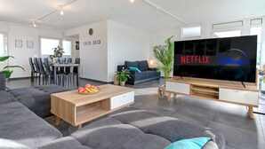 A modern living room with a grey sectional sofa, a wooden coffee table with fruit, a TV displaying Netflix and Disney+, and a dining area with chairs and a table.