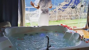 A person in a white robe standing by a window with a mountain view, a hot tub with rose petals, and a sign indicating a temperature of 38 degrees Celsius. - Nirvana Hamta Retreat (Manali)