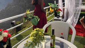 A woman in a red dress standing at a glass table with a bottle of wine, surrounded by greenery and a mountainous backdrop. - Nirvana Hamta Retreat (Manali)
