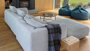 A modern living room with a grey sectional sofa, a small round coffee table, and a fireplace. There are large windows offering a view of a green landscape. - Te Whare Waipara

 (Broomfield)