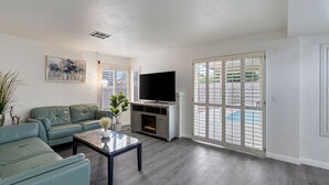 A living room with a green sofa, a flat-screen TV, a fireplace, and a large sliding glass door leading to a pool.