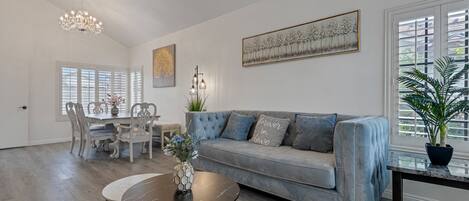 A spacious living room with a grey sofa, a dining table with chairs, a chandelier, and a large window with blinds.