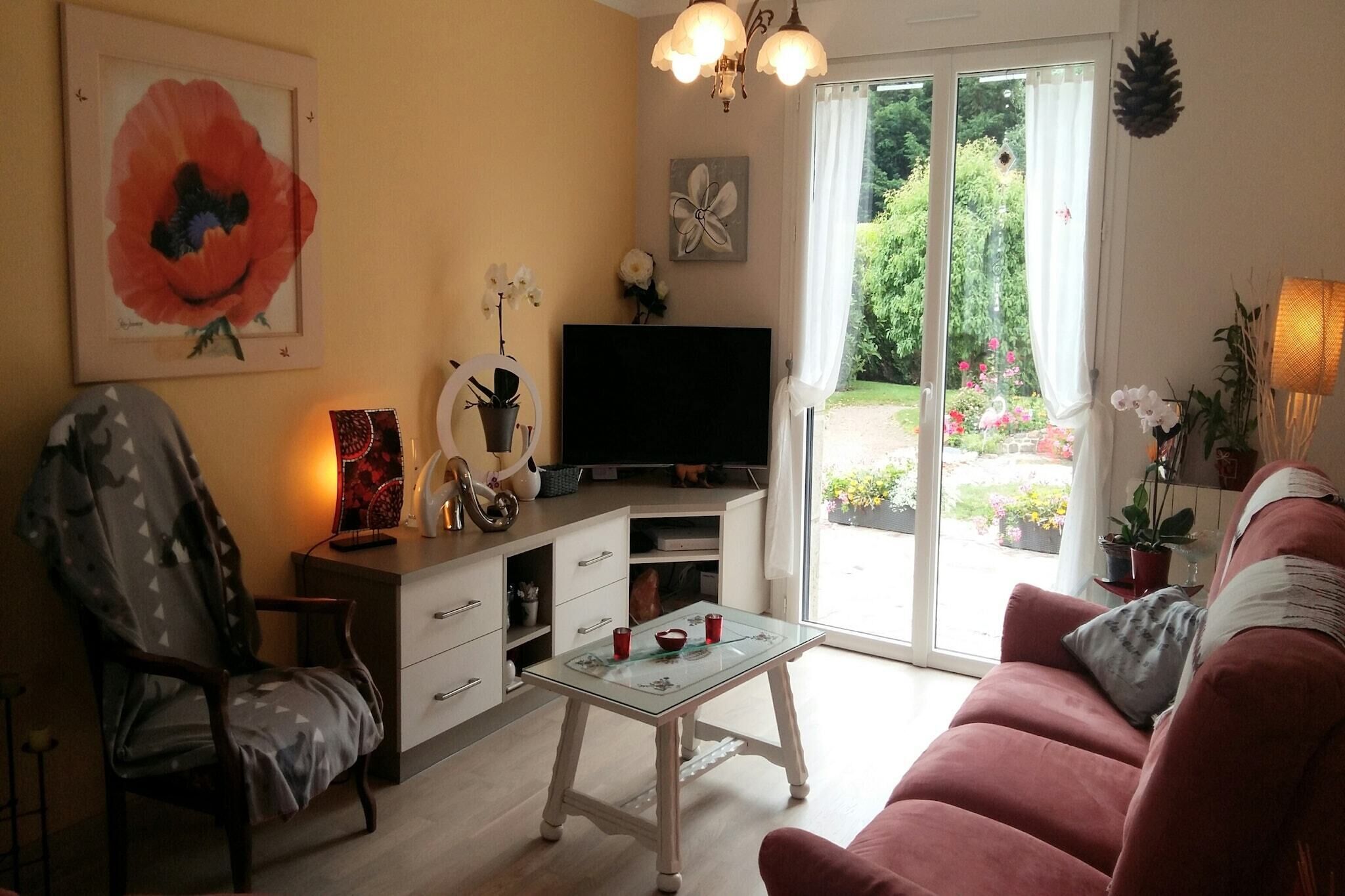 A living room with a sofa, a chair, a glass coffee table, a flat-screen TV, and a sliding glass door leading to a garden.