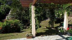 Un patio abrité doté d’une structure en forme de pergola, entouré d’une végétation luxuriante et de plantes à fleurs.
