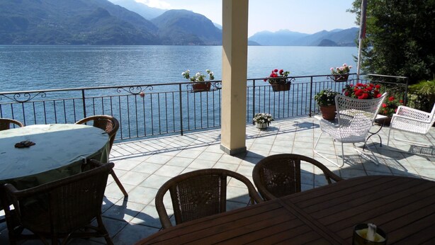Une terrasse offrant une vue sur un lac et des montagnes, avec des chaises en osier, une table et des plantes en pot.