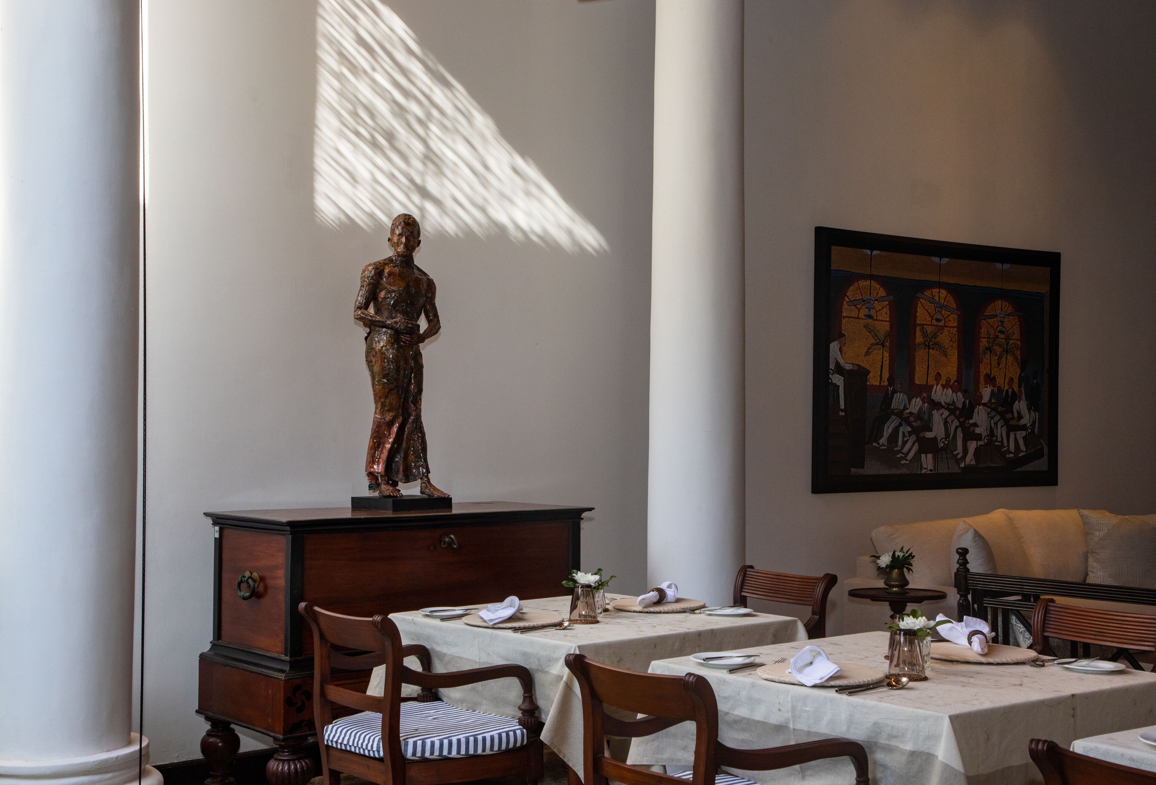 Un comedor con una estatua sobre un aparador, mesas dispuestas para comer y un cuadro en la pared.