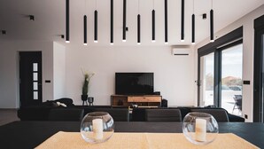 A modern living room with a black sofa, a TV on a wooden stand, and a dining table with two glass candle holders.