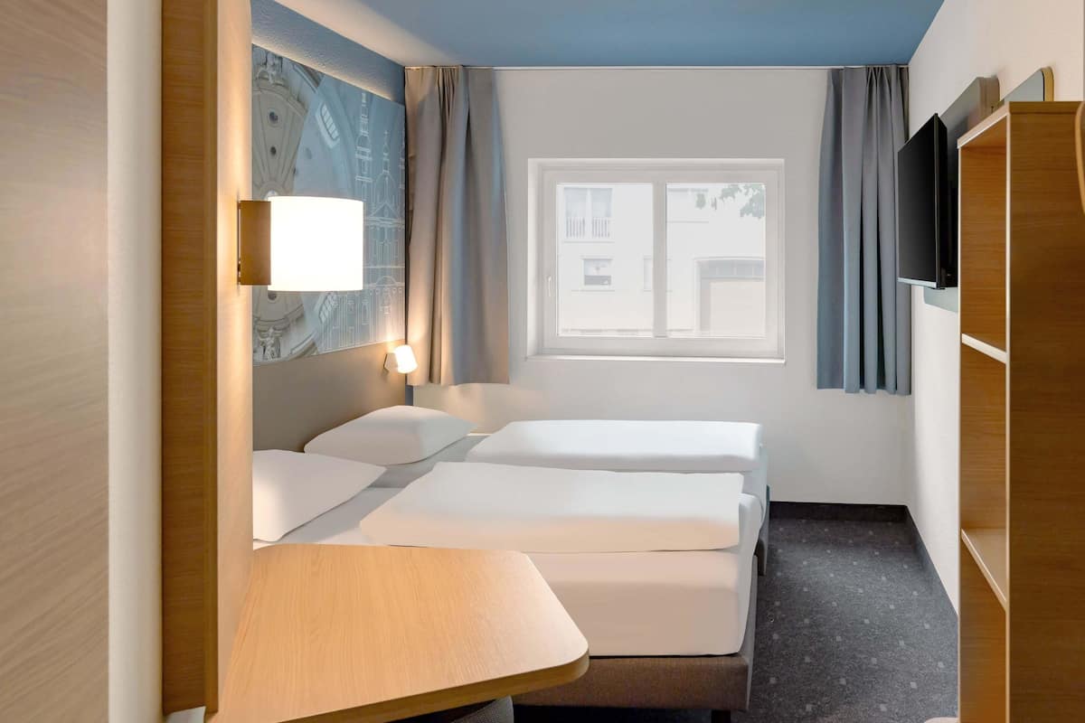 A hotel room with two beds, a desk, a TV, and a window with curtains.