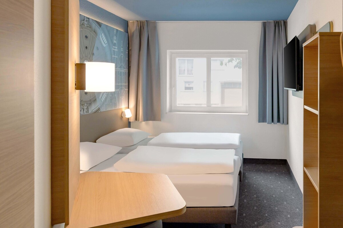A hotel room with two beds, a desk, a TV, and a window with curtains.