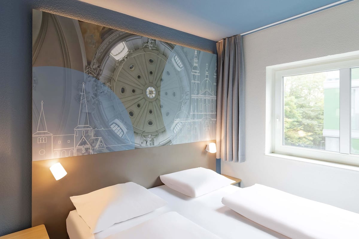 A hotel room with a large wall mural of a clock tower, a bed with white linens, a bedside table, and a window with curtains.