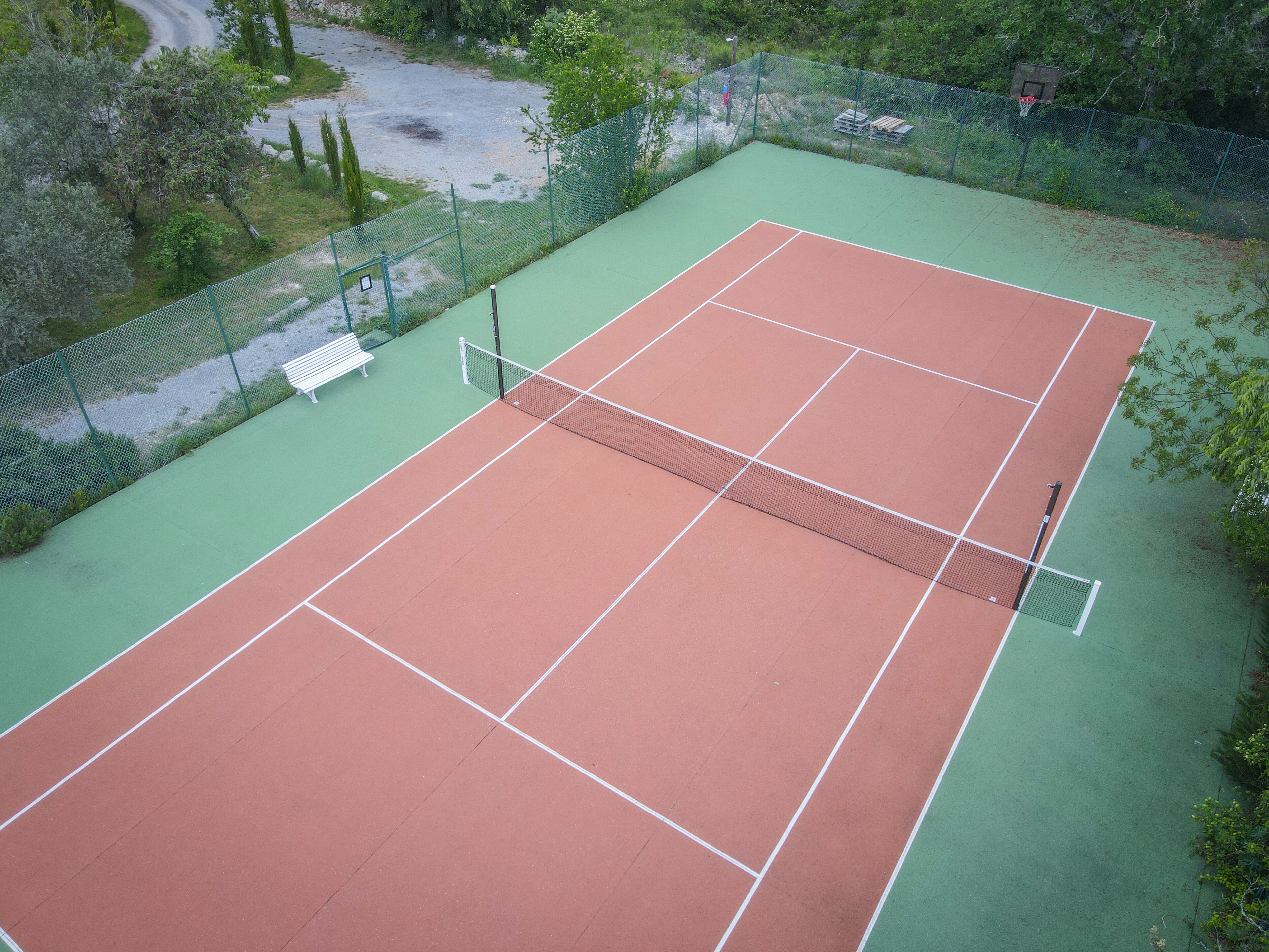 An outdoor tennis court with a green and red surface, surrounded by a fence and trees.