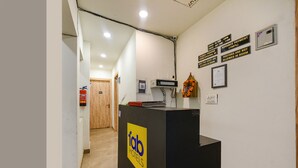A hotel reception area with a reception desk, a fire extinguisher, and a wooden door in the background. - FabExpress Divya Villa (New Delhi)