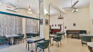 A dining area with tables and chairs, a piano, and ceiling fans. - Fabhotel Sarovar Palace (Prayagraj)