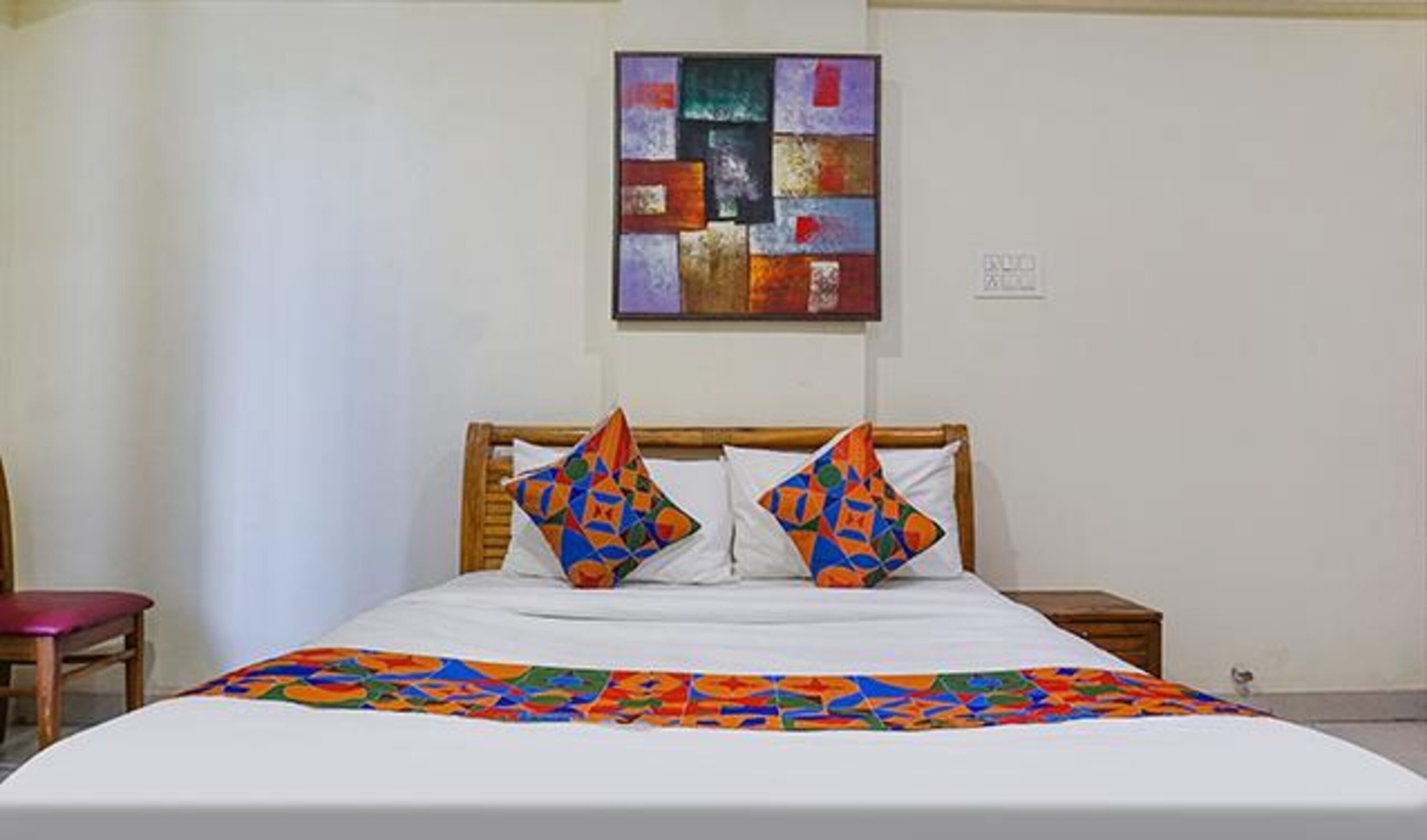 Een bed met een kleurrijke quilt, twee kussens met geometrische patronen, en een meerkleurige kunstwerk erboven.