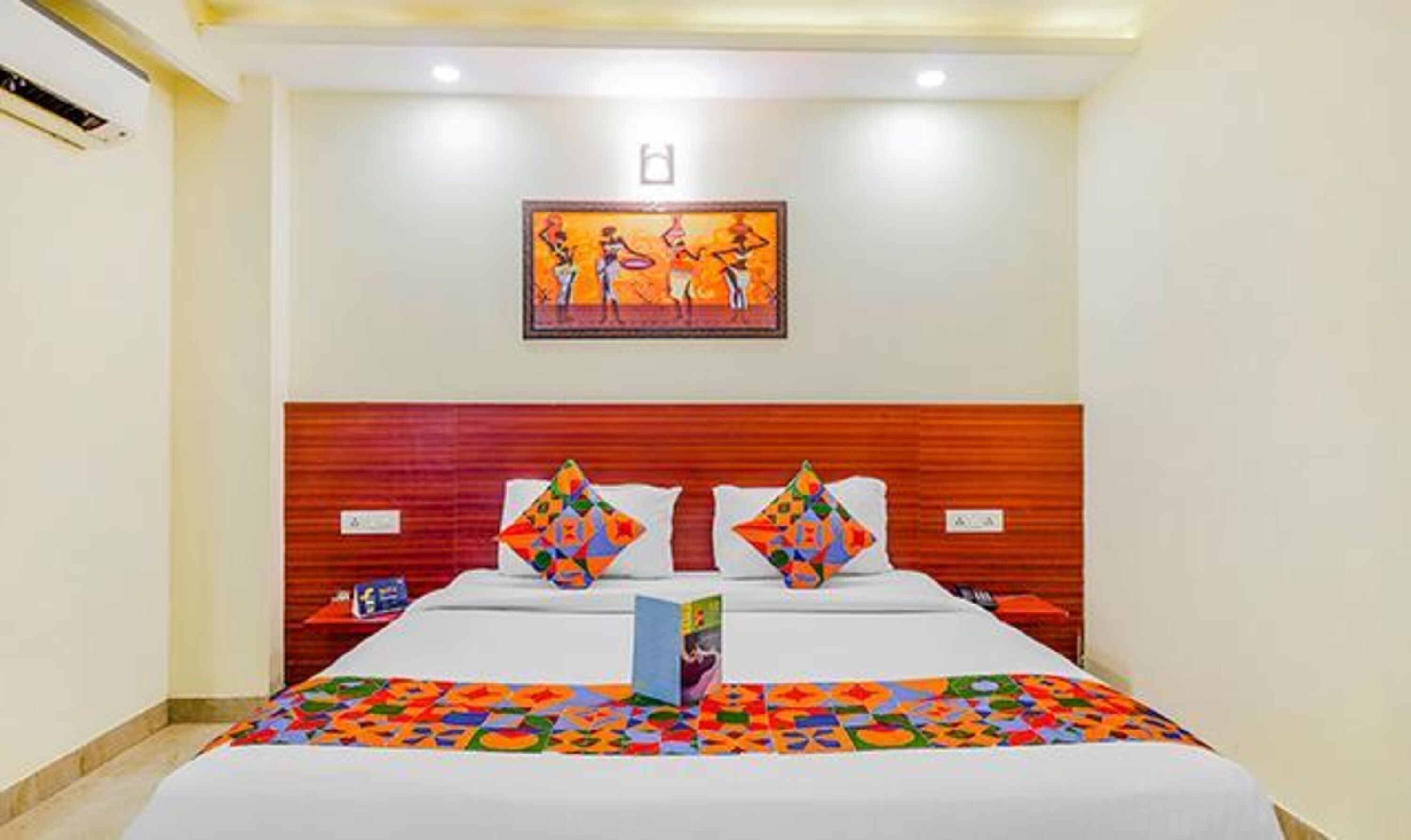 A hotel room with a large bed, two pillows, a nightstand, a wall-mounted painting, and a ceiling light.