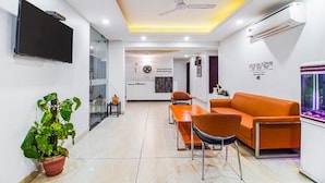A modern hotel lobby with a flat-screen TV, orange seating, a potted plant, and a fish tank.