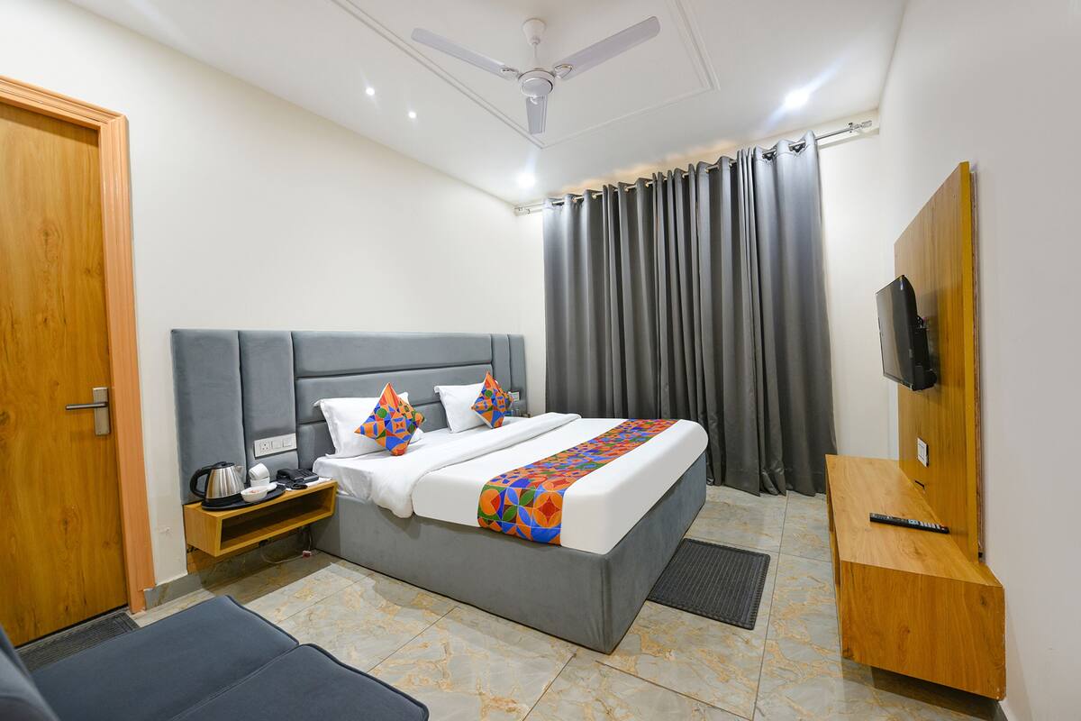A hotel room with a bed, a TV, a wooden desk, a ceiling fan, and a window with curtains.
