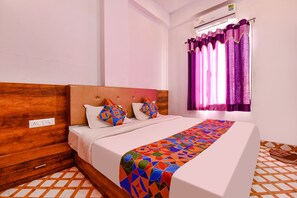 A neatly made bed with a colorful geometric-patterned bedspread, a wooden headboard, and a white wall with a power outlet. - Fabexpress Raj Palace (Udaipur)