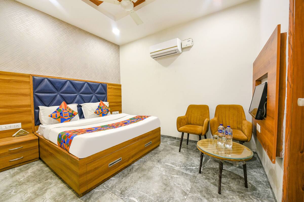A hotel room with a wooden bed, two armchairs, a small table with water bottles, an air conditioning unit, and a TV mounted on the wall.