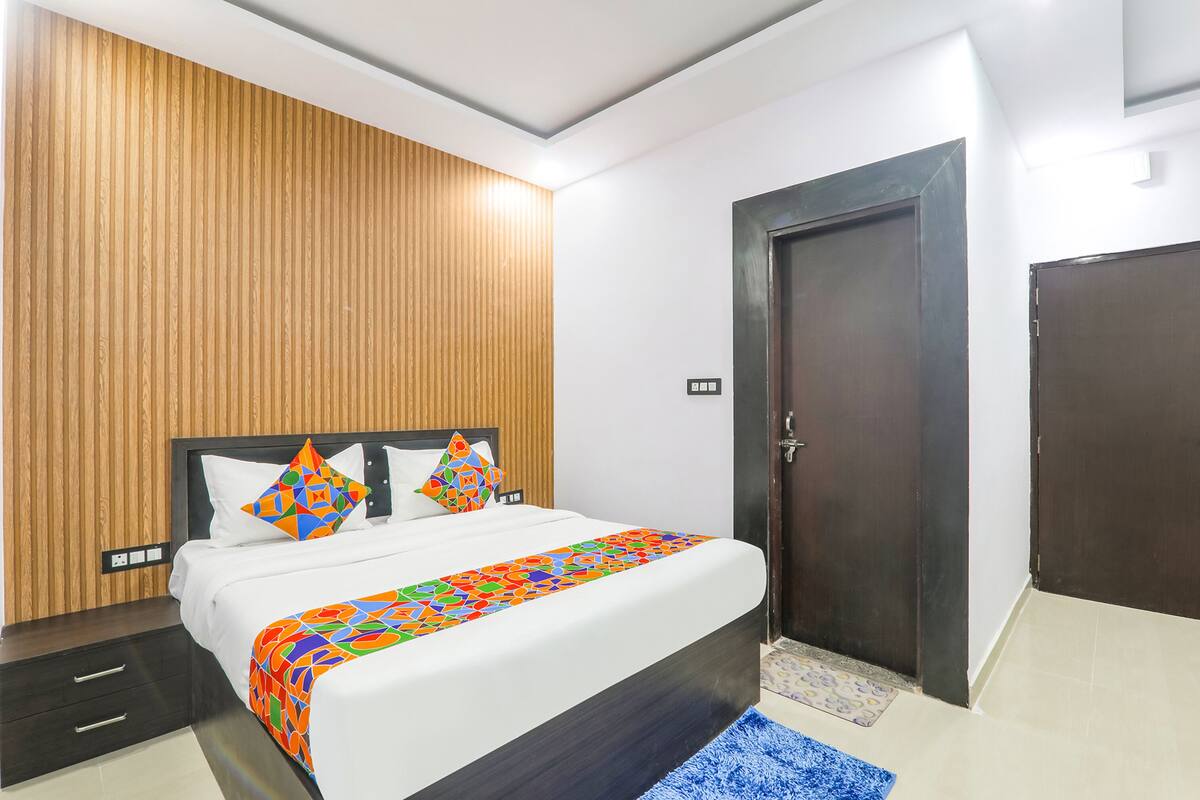 A bedroom with a wooden panel wall, a bed with white bedding and colorful pillows, a dark door, and a blue rug.