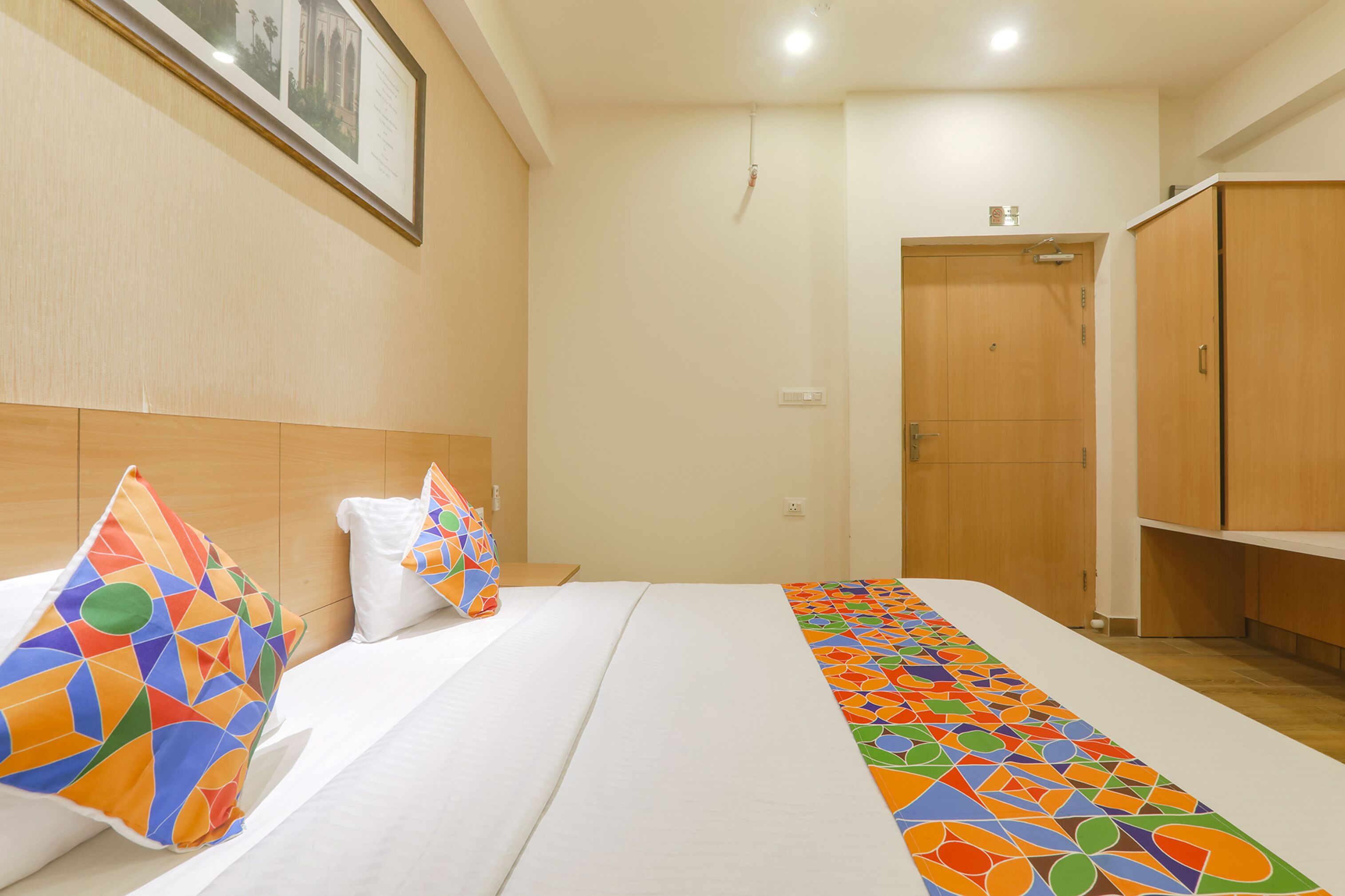 A hotel room with a bed, a colorful pillow, a framed picture on the wall, and a wooden wardrobe.