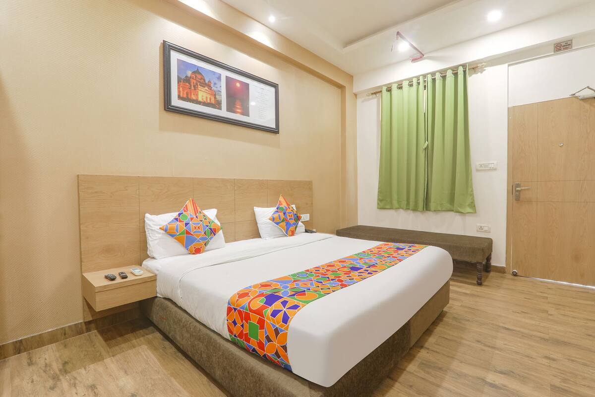 Habitación de hotel con una cama grande, cabecera de madera, mesita de noche con teléfono y control remoto, un banco, cortina verde y un cuadro en la pared.