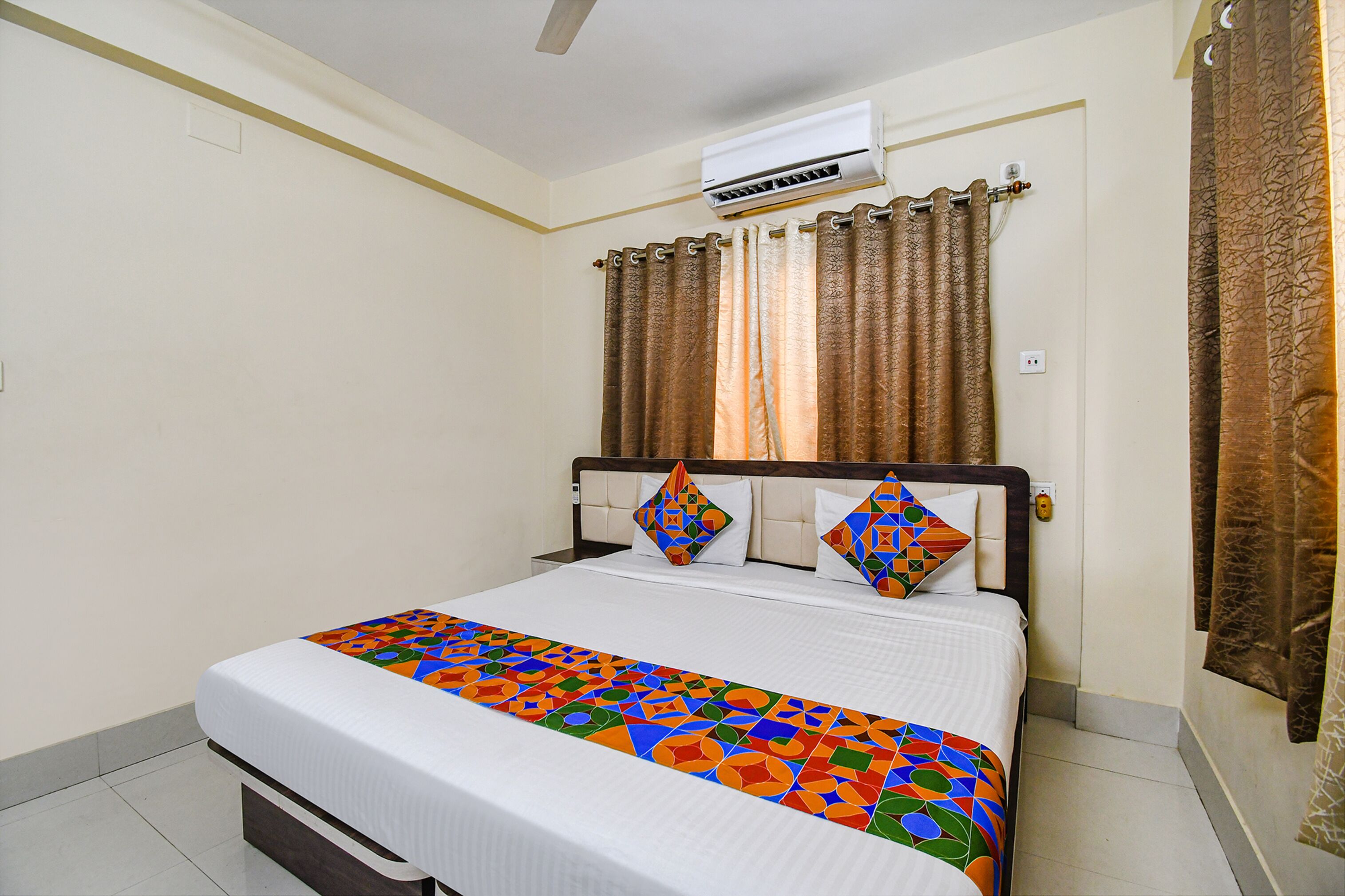 A hotel room with a bed, an air conditioning unit, curtains, and a patterned bedspread.