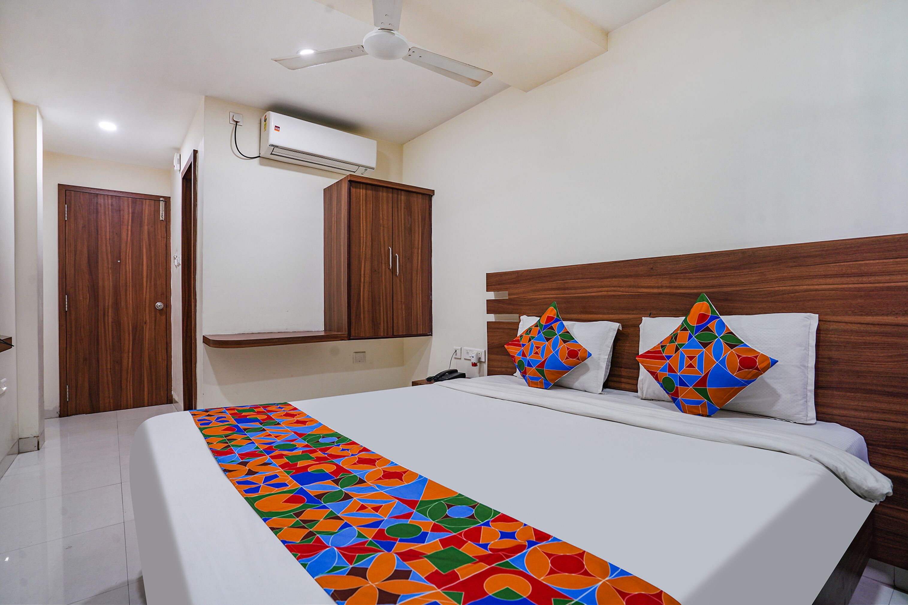 Ein Hotelzimmer mit Bett, Deckenventilator, Klimaanlage, Holzschrank und einer farbenfrohen Bettdecke.